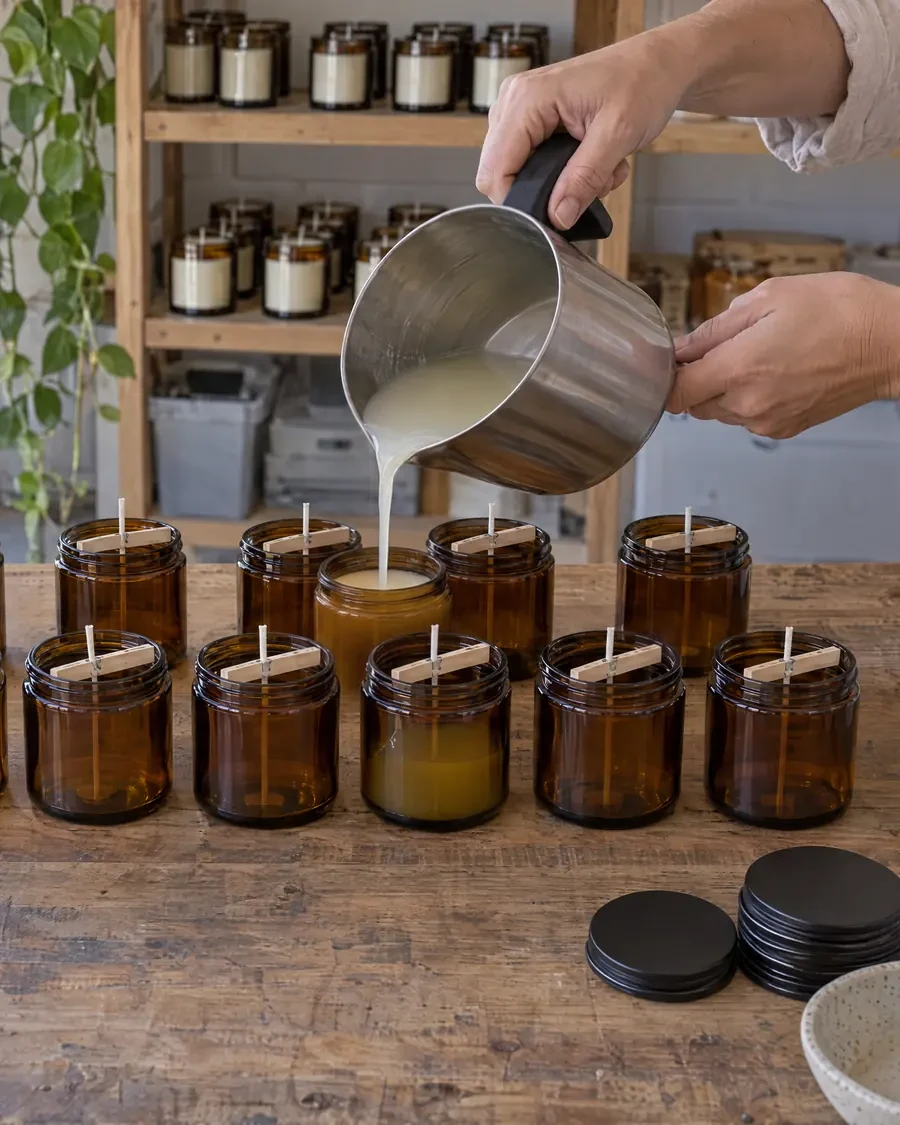 Twelve amber jars with wicks on a hardwood bench, wax being poured into the front row